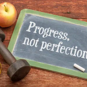 Image of blackboard with dumbells and apple. Written on the blackboard is "progress, not perfection"