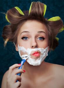 woman shaving her face with curlers in her hair. PCOS hormonal acne is linked to high androgens that also cause facial hair in women