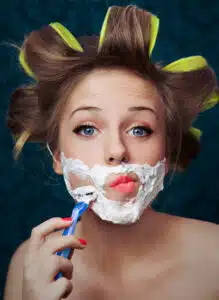 woman shaving her face with curlers in her hair. PCOS hormonal acne is linked to high androgens that also cause facial hair in women